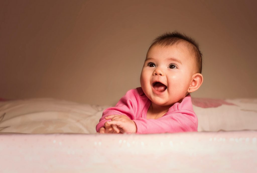 Lächelndes Baby in rosa Strampler liegt auf dem Bett und schaut begeistert nach oben – Symbolbild für zufriedenes Baby nach Fläschchen mit baby flaschenzubereiter.