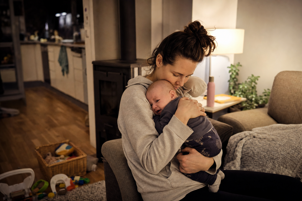 Ein Elternteil beruhigt am Abend ein unruhiges Baby in einem warm beleuchteten Wohnzimmer.
