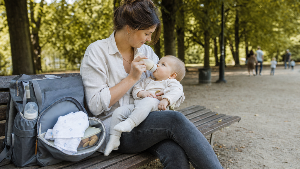 Mutter fuettert ihr Baby unterwegs auf einer Parkbank, mit Wickelrucksack, Flasche und Snacks griffbereit