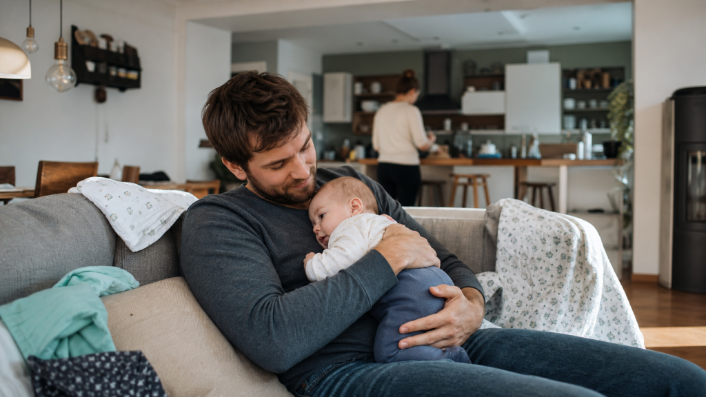 Elternteil mit Baby im Arm in einer gemütlichen, leicht unordentlichen Wohnung als realistischer Familienalltag mit Baby